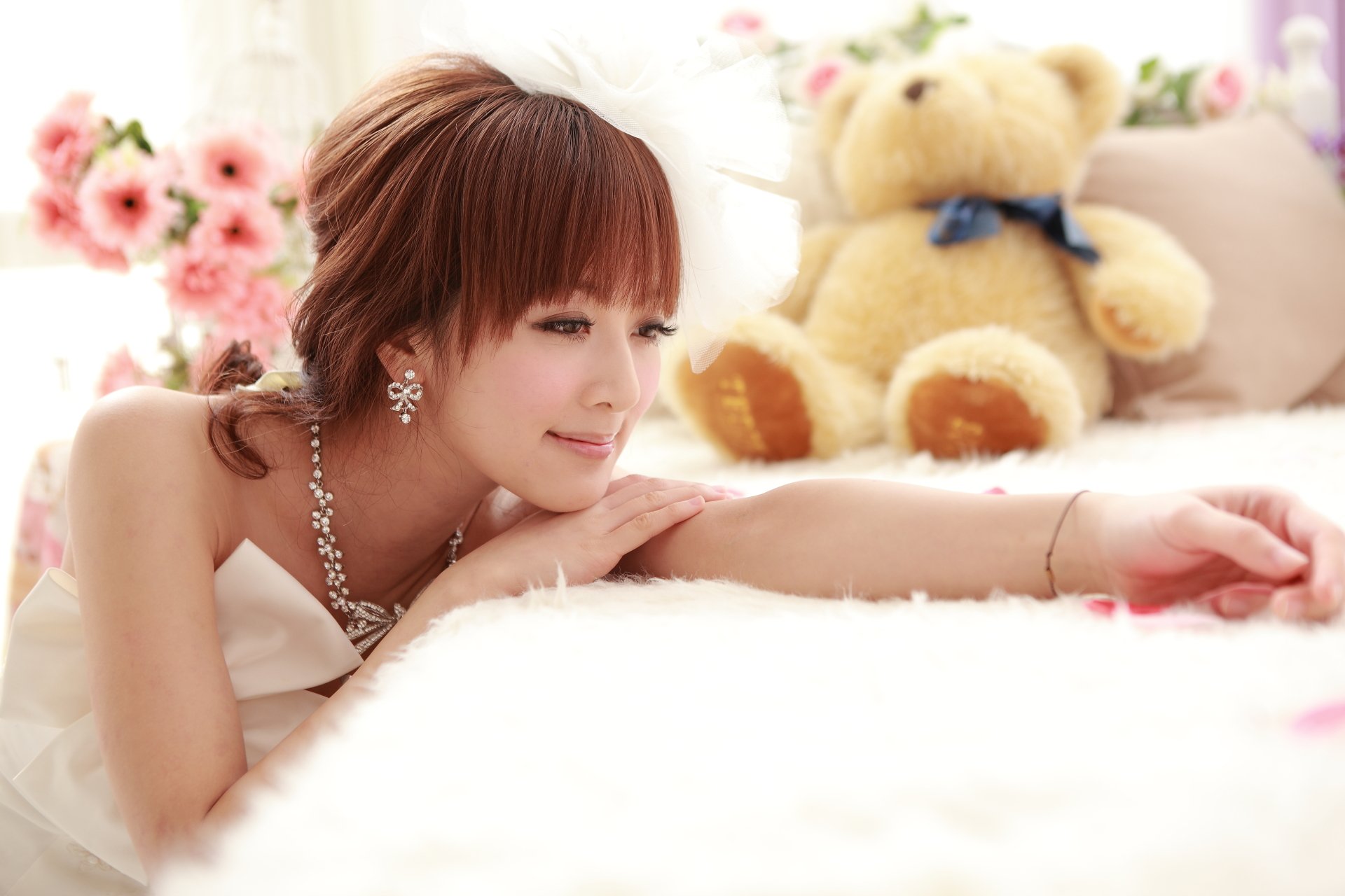 A Taiwanese woman in a white dress smiles while lying on a soft surface, with a teddy bear and flowers in the background. This HD wallpaper features Mikako Zhang Kaijie.