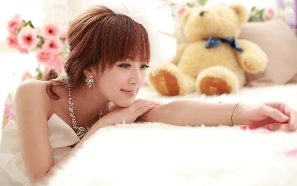 A Taiwanese woman in a white dress smiles while lying on a soft surface, with a teddy bear and flowers in the background. This HD wallpaper features Mikako Zhang Kaijie.
