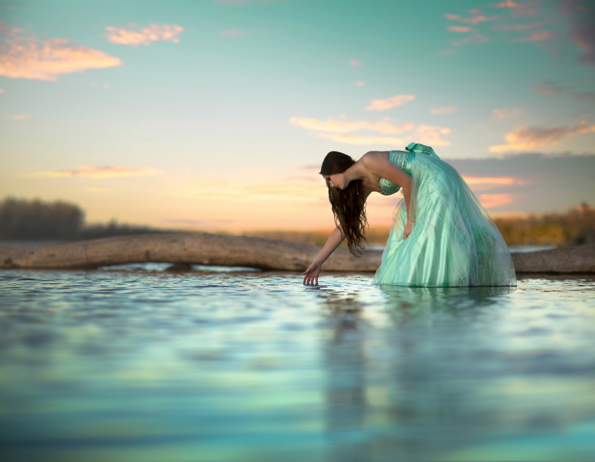 HD desktop wallpaper featuring a serene portrait of a woman in a flowing teal dress bending over water at sunset, capturing a calm and reflective mood.