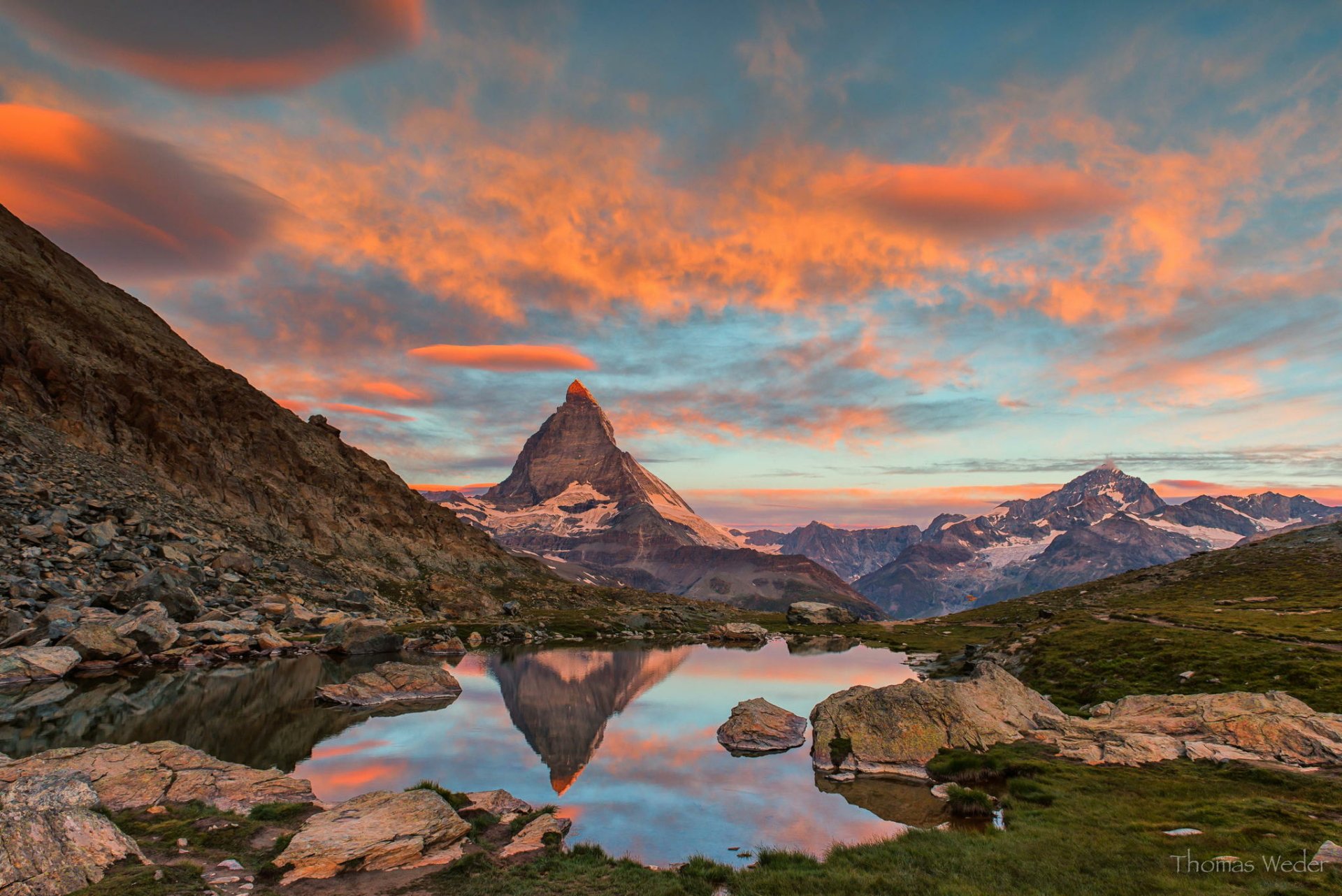 HD desktop wallpaper: the Matterhorn rising beneath glowing clouds and sky, mirrored in a mountain tarn—rugged alpine nature with a clear reflection.