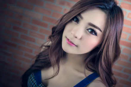 HD portrait of an Asian Thai woman with long, wavy hair and a gentle expression, set against a brick wall background. The tag 'Kookai' is featured.