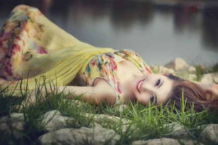 An Asian woman in a colorful Thai dress is smiling while lying on grass and rocks, with a serene water background. HD desktop wallpaper.