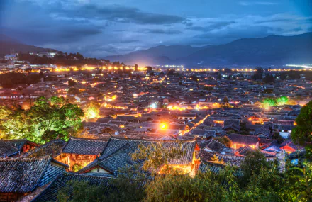 A 4K Ultra HD view of Lijiang village in Yunnan, China, showcasing illuminated rooftops under a cloudy sky with mountains in the background.