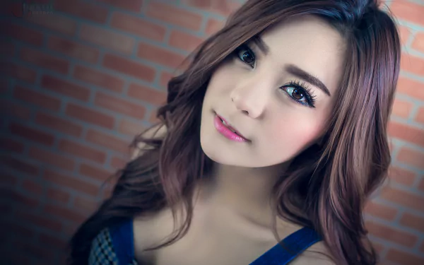 HD portrait of an Asian Thai woman with long, wavy hair and a gentle expression, set against a brick wall background. The tag 'Kookai' is featured.