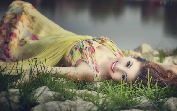 An Asian woman in a colorful Thai dress is smiling while lying on grass and rocks, with a serene water background. HD desktop wallpaper.