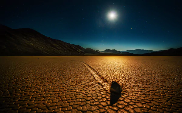 A stunning night sky above California's Death Valley, featuring a glowing moon and a solitary stone leaving a trail on the cracked desert floor, captured in 4K Ultra HD.