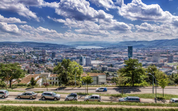 5K Ultra HD desktop wallpaper of Zurich, Switzerland: panoramic cityscape and lake beneath dramatic clouds; foreground road with parked cars and urban greenery highlighting man-made structures.