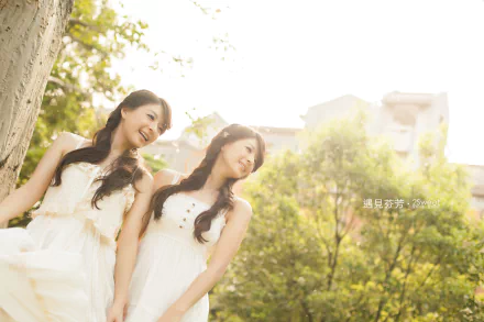 Two smiling women in white dresses leaning against a tree in a sunlit park with soft bokeh — 5K Ultra HD PC desktop wallpaper and background.