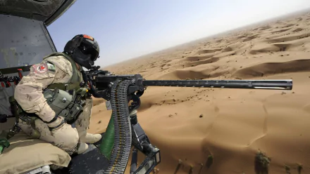 A soldier in tactical gear operates a machine gun from a helicopter, overlooking vast desert landscapes. This high-definition image captures a moment of military readiness.