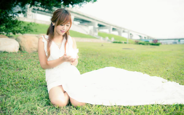Taiwanese model Xiao Xi in a white dress kneeling on grass near a bridge, captured in 4K Ultra HD for a PC desktop wallpaper and background.