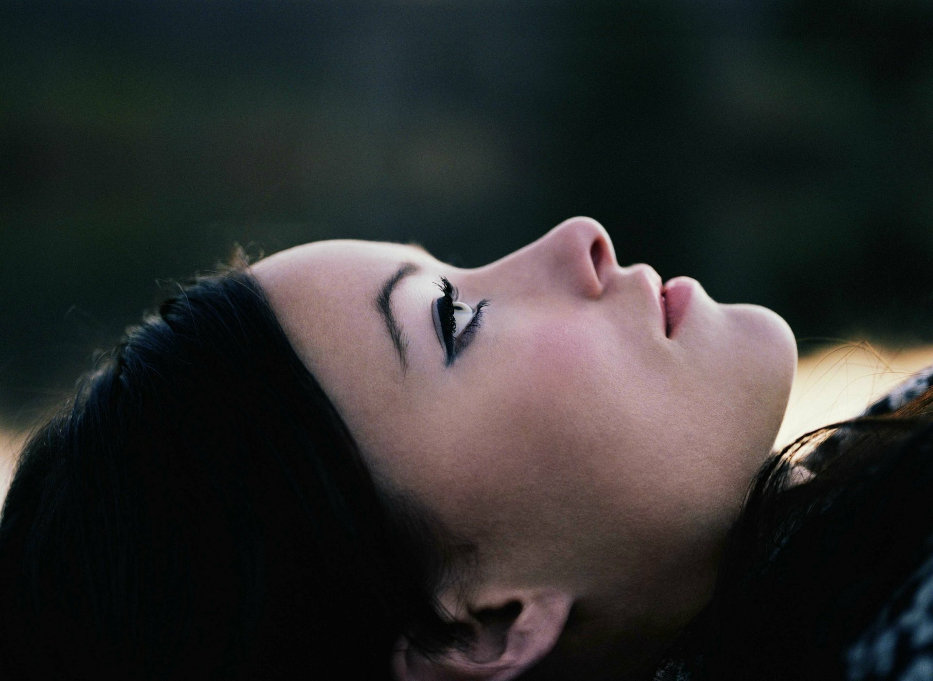 Close-up profile of a reclining woman with dark hair and winged eyeliner against a soft blurred background — 2K Quad HD PC desktop wallpaper.