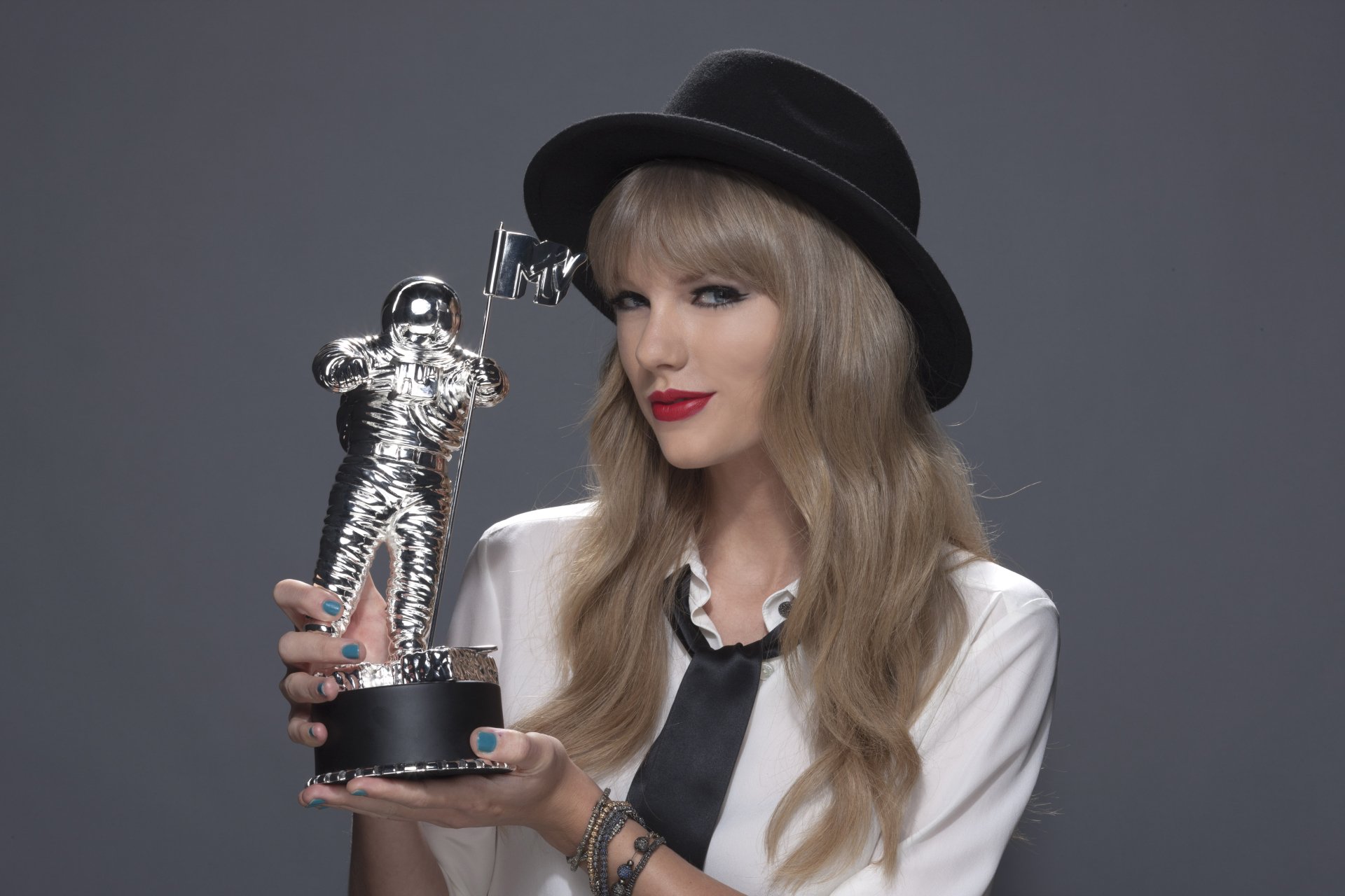 Taylor Swift, an American singer with blonde hair and red lipstick, wears a stylish hat while holding a silver award statue. The image captures her confident, iconic presence.