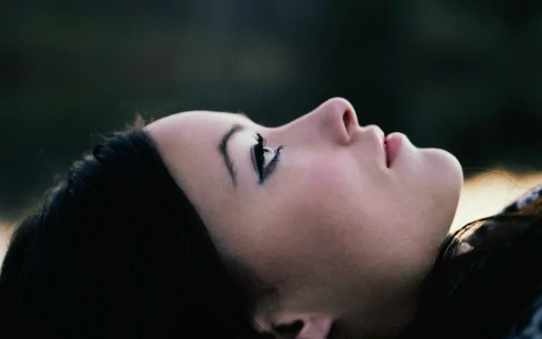 Close-up profile of a reclining woman with dark hair and winged eyeliner against a soft blurred background — 2K Quad HD PC desktop wallpaper.