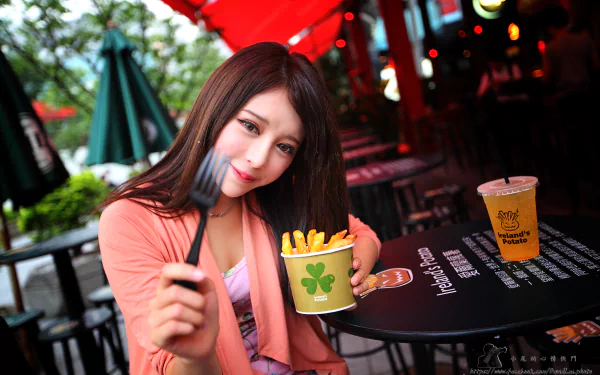 A Taiwanese model, Julie Chang, smiles while holding a fork and a cup of fries at a vibrant outdoor setting, with drinks on the table, captured in stunning 4K Ultra HD.