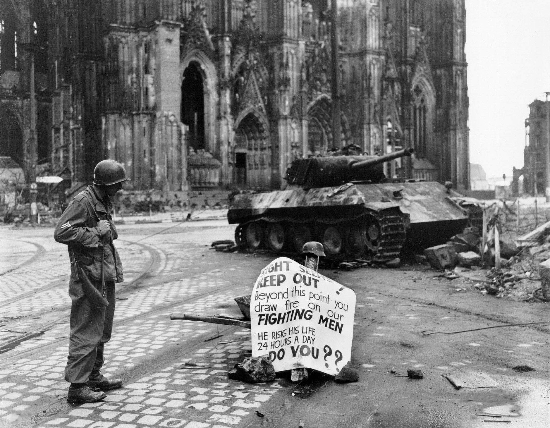 Black and white HD desktop wallpaper showing a soldier and a tank in a war-torn urban setting with a sign questioning the fighting men, evoking military themes.