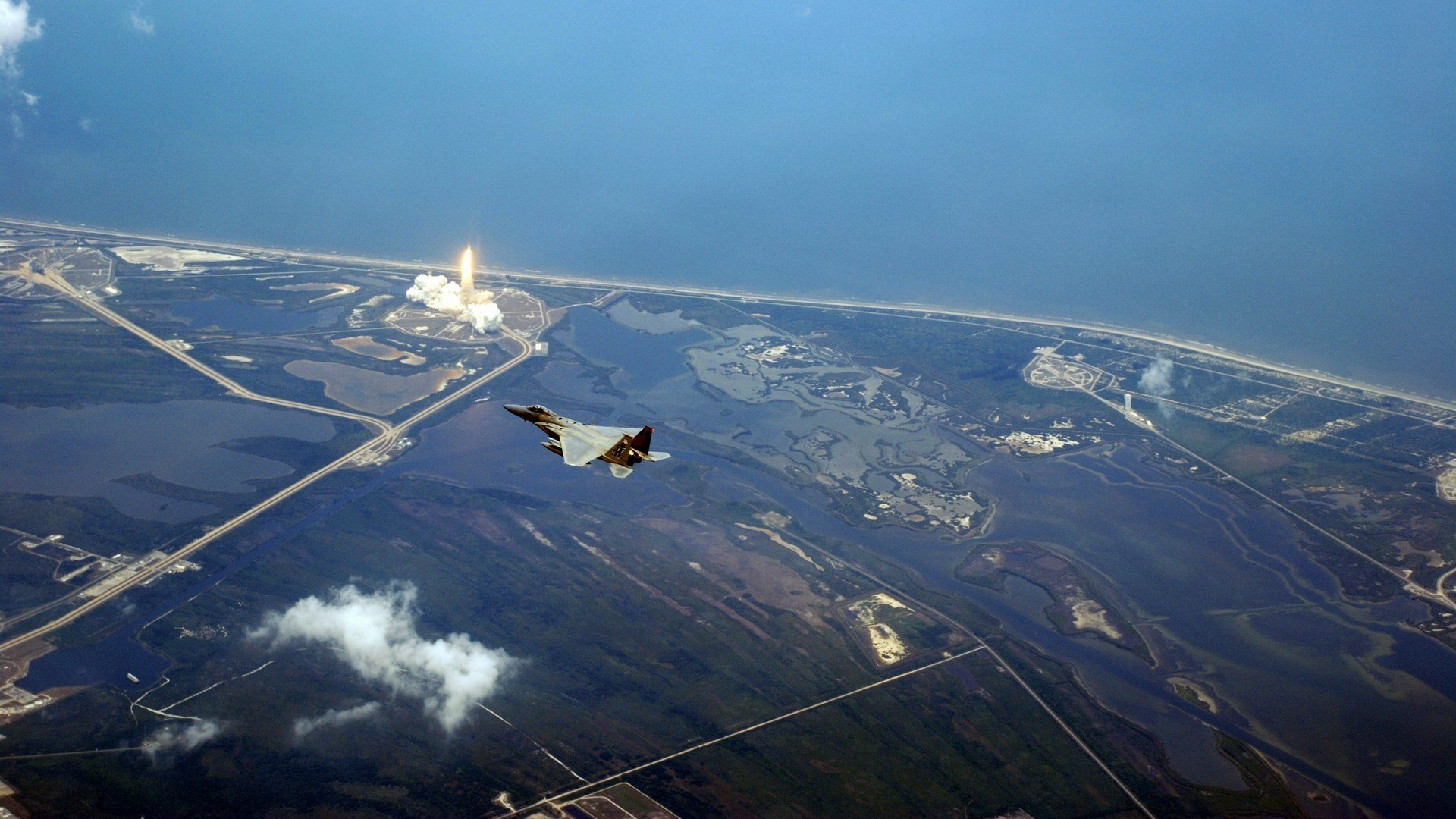 HD PC desktop wallpaper/background of a military McDonnell Douglas F-15 Eagle flying over coastal landscape with facilities visible below.