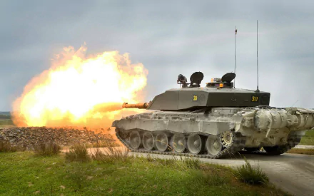 Military Challenger 2 tank firing its main gun across open terrain — HD PC desktop wallpaper/background with dramatic muzzle flash and cloudy sky.