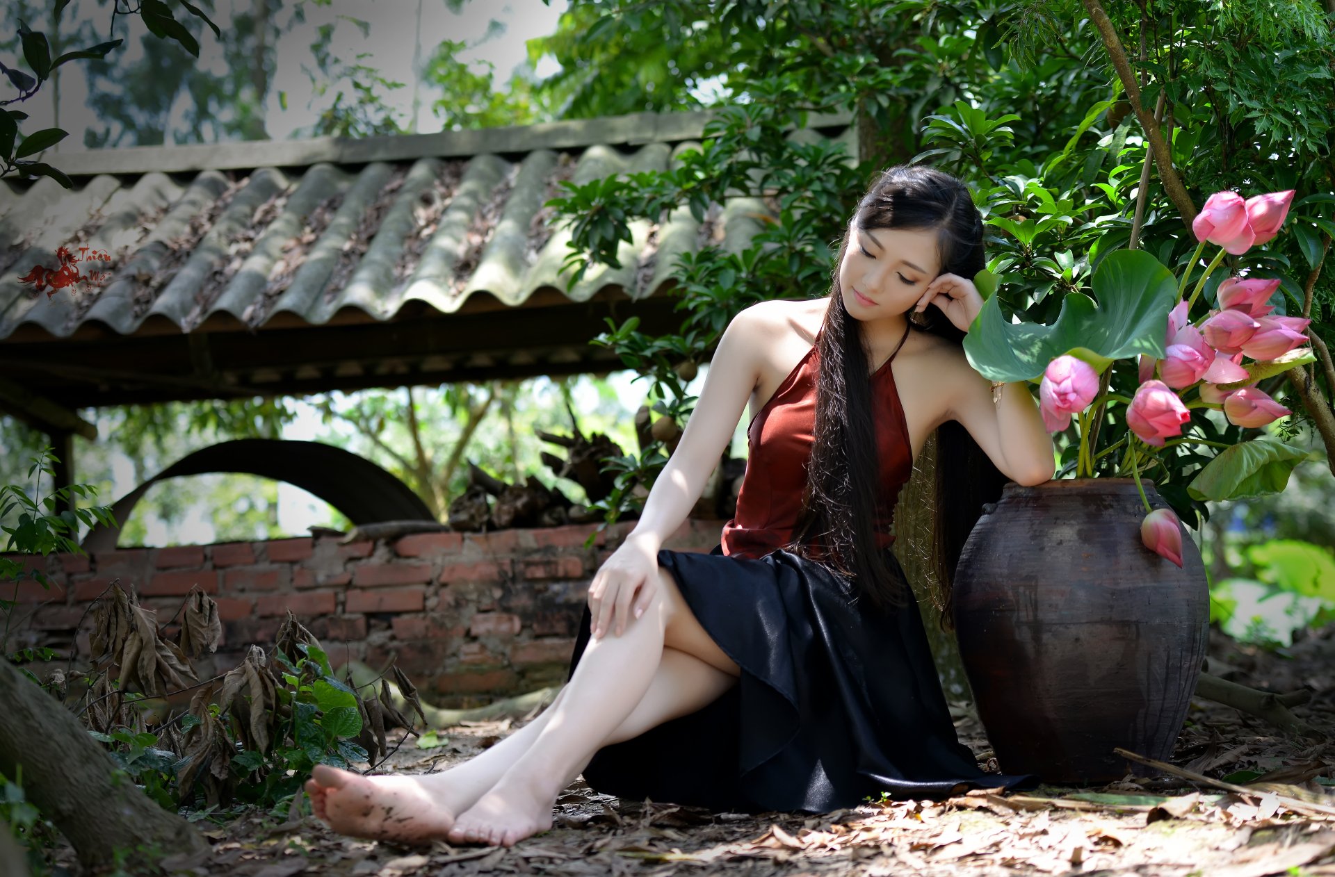 A Vietnamese woman in a traditional outfit sits barefoot beside a large vase with pink lotus flowers in a serene, outdoor setting. The HD desktop wallpaper showcases nature and tranquility.