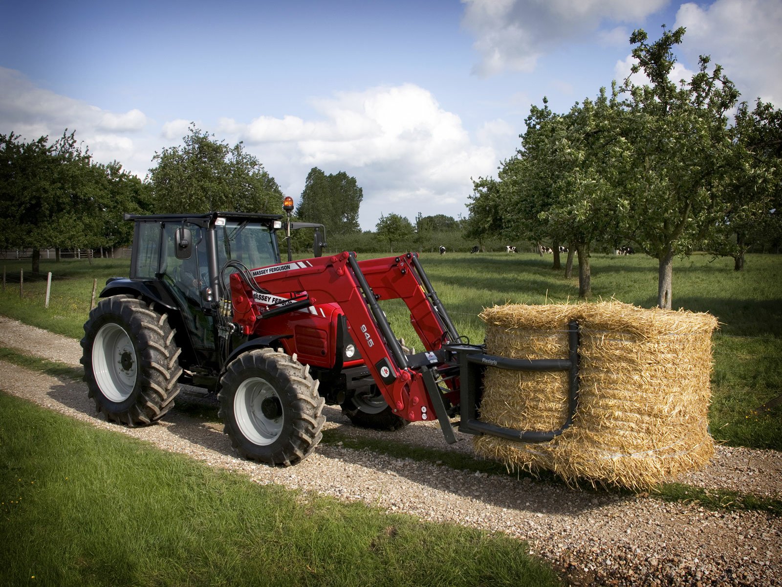 A Massey Ferguson tractor rolls down a gravel path, transporting a large round hay bale amid lush green trees, creating a vibrant rural scene in this HD desktop wallpaper.