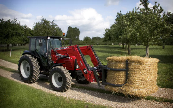 A Massey Ferguson tractor rolls down a gravel path, transporting a large round hay bale amid lush green trees, creating a vibrant rural scene in this HD desktop wallpaper.