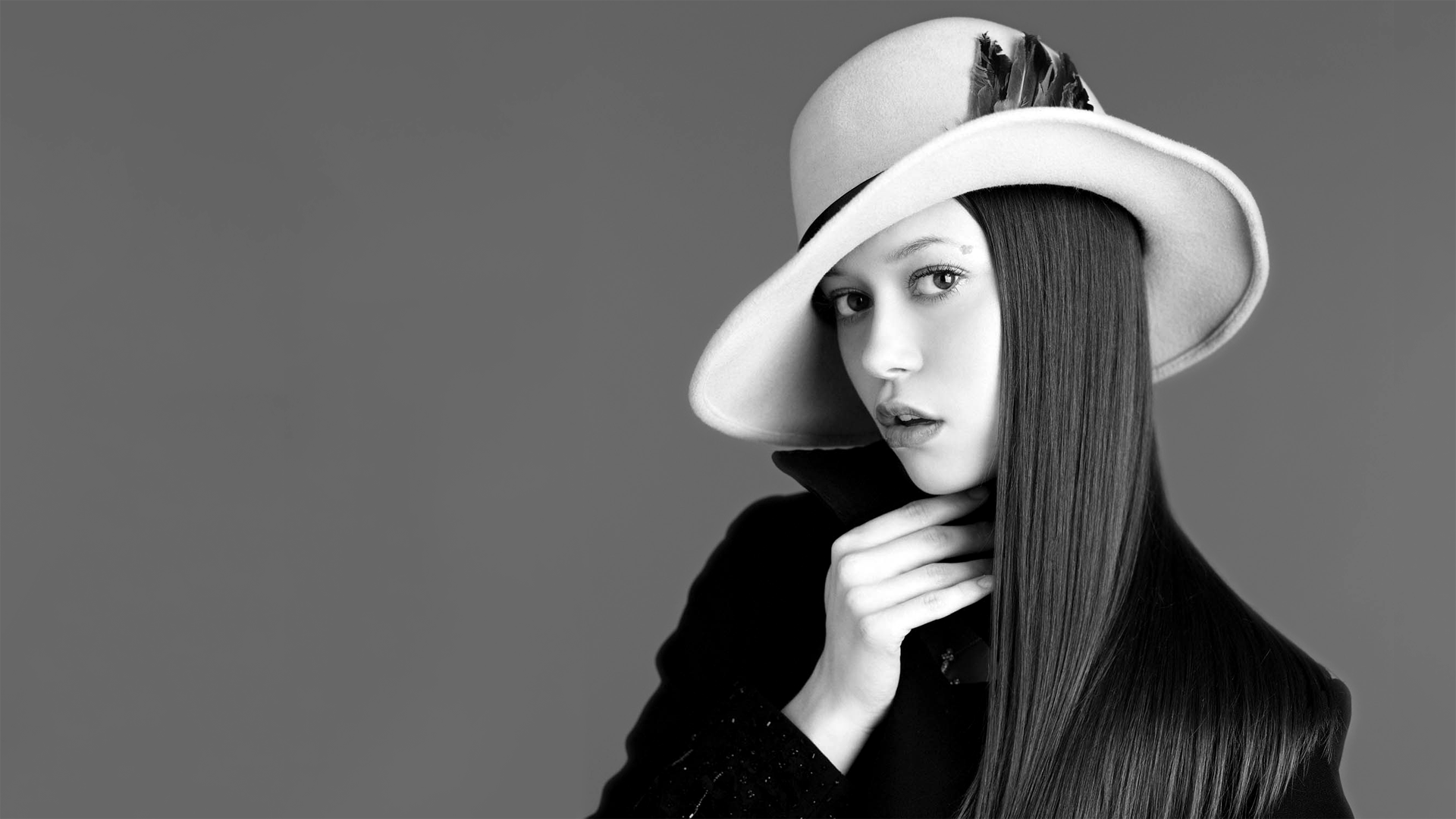 Black-and-white HD PC desktop wallpaper of a female celebrity in a wide-brim hat and dark coat, long straight hair, hand at her throat, looking directly at the camera.