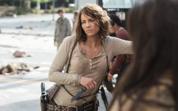 HD desktop wallpaper featuring Lauren Cohan as Maggie Greene in a scene from the TV show The Walking Dead. Maggie is standing outdoors, armed with a gun and knife, amidst a post-apocalyptic setting.
