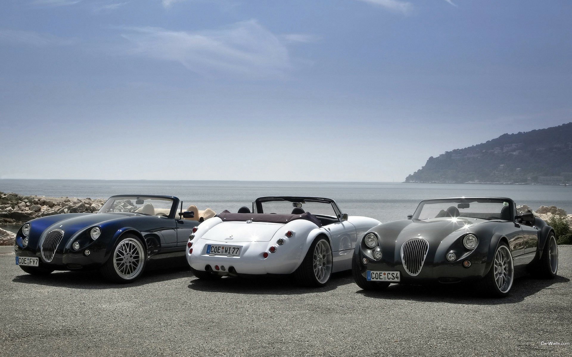 HD PC desktop wallpaper and background showing three Wiesmann vehicles — black, white and gray roadsters parked by the seaside beneath a clear sky.