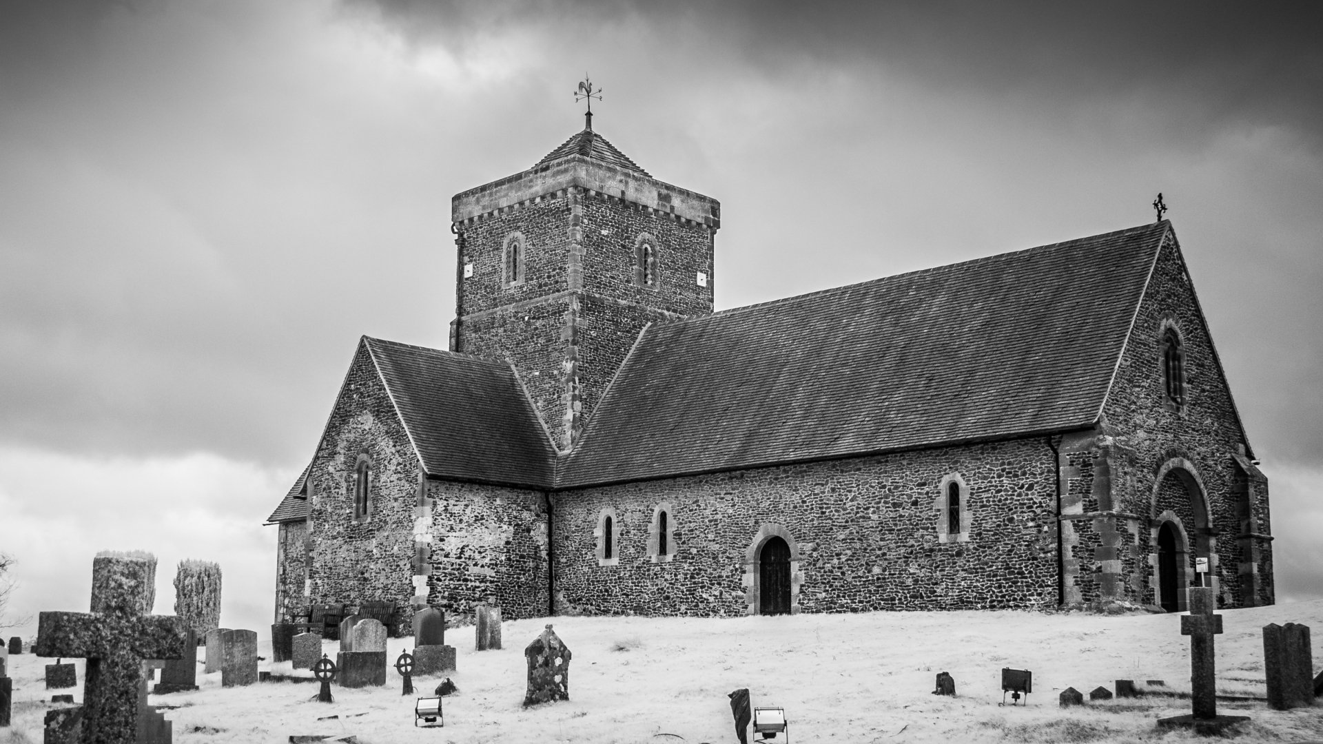 Black and white HD wallpaper of the Church of St Martha-on-the-Hill, a historic religious site in Surrey, England, with surrounding gravestones under a cloudy sky.