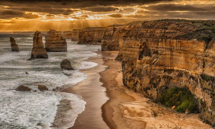 HD desktop wallpaper of The Twelve Apostles limestone stacks along the rugged coastline of Melbourne, Victoria, Australia, under a dramatic golden sunset sky.