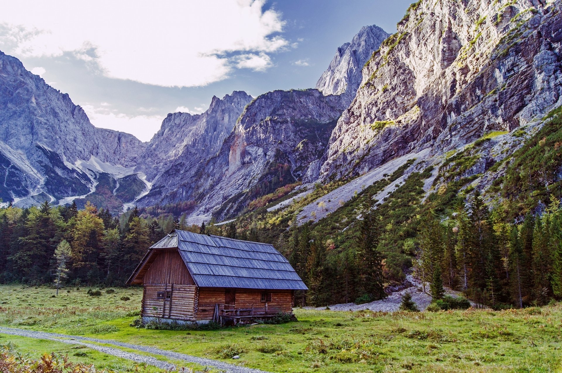 4K Ultra HD PC desktop wallpaper: landscape of a wooden hut beside a tree in a green countryside meadow, pine forest at the foot of towering rocky mountains under a bright sky.