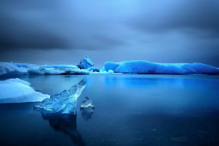 HD PC desktop wallpaper/background depicting a serene nature scene: glowing blue icebergs reflected on calm Arctic waters beneath a moody, cloud-filled sky.