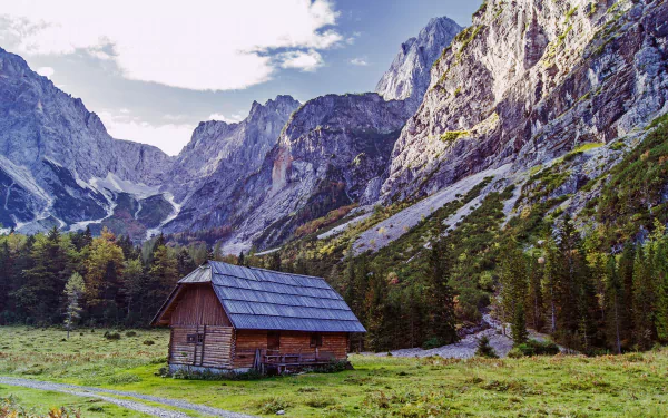 4K Ultra HD PC desktop wallpaper: landscape of a wooden hut beside a tree in a green countryside meadow, pine forest at the foot of towering rocky mountains under a bright sky.