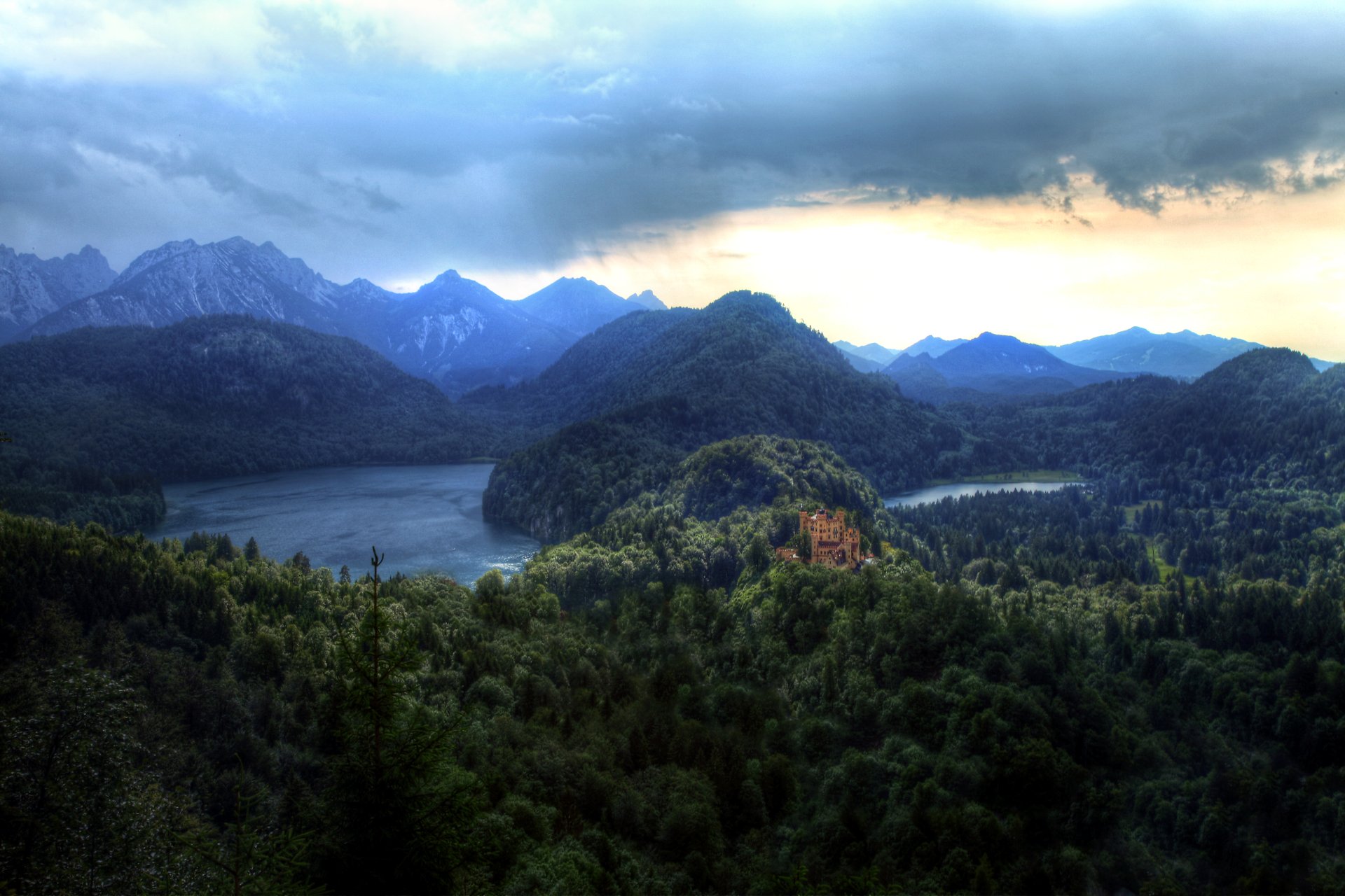 Hohenschwangau Castle above a forested lake in Gemeinde Schwangau, Bavaria, with cloud-lit Alps and mountains in the background — dramatic 5K Ultra HD desktop landscape.