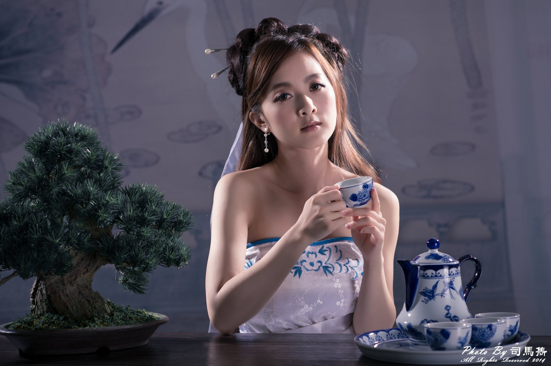 A Taiwanese woman dressed in traditional Chinese attire with a hairpin, holding a tea cup beside a bonsai and a blue porcelain tea set, captured in 4K Ultra HD.