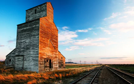 HD countryside landscape of a worn train station beside railroad tracks, set against a blue sky with scattered clouds.