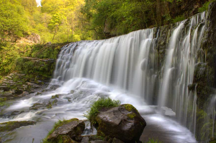 Sgwd Isaf Clun-gwyn Waterfall cascading over rocks surrounded by lush green foliage in Ystradfellte, South Wales, Neath, United Kingdom. 4K Ultra HD landscape wallpaper.