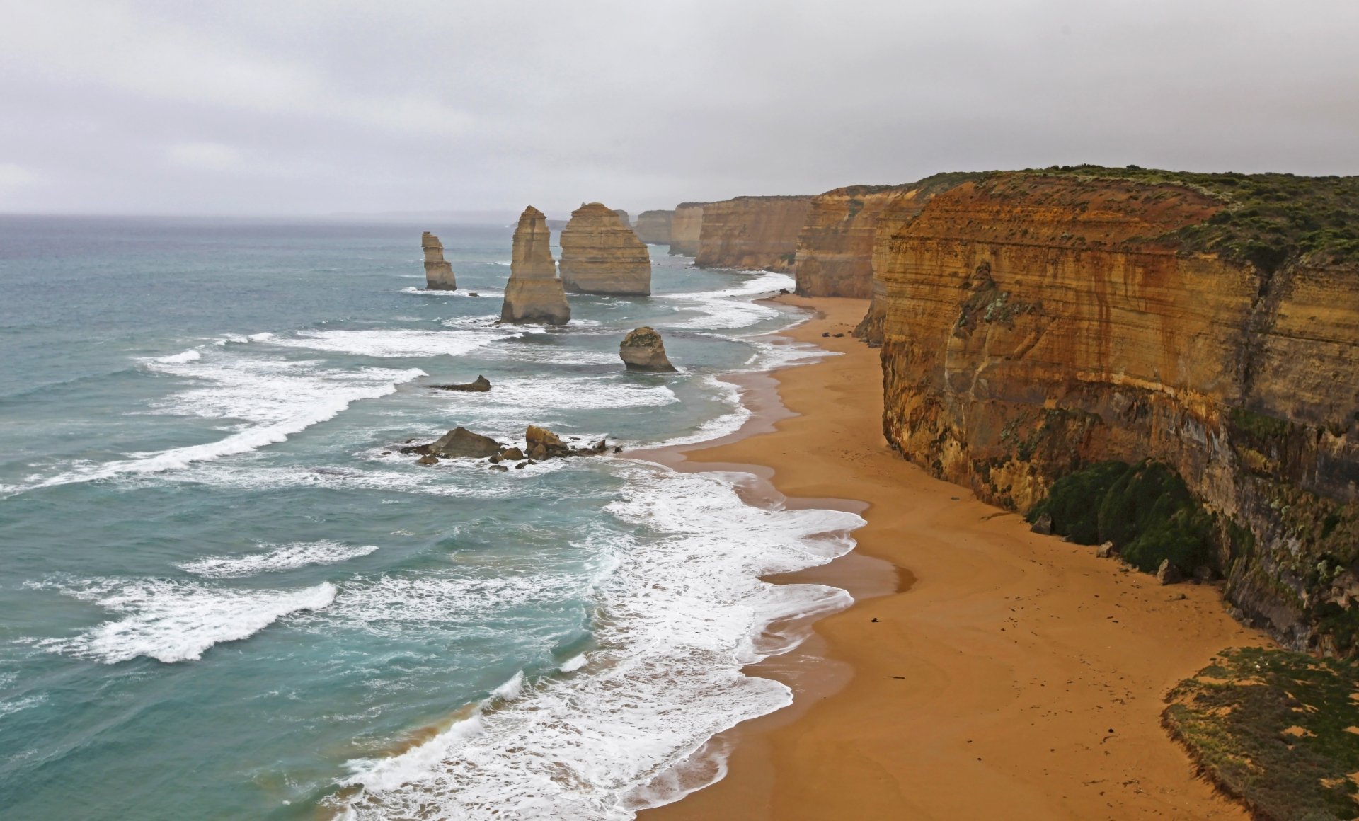 Waves crash along the sandy coastline of Victoria, Australia, showcasing the limestone stacks of The Twelve Apostles against a cloudy sky in this 4K Ultra HD wallpaper.