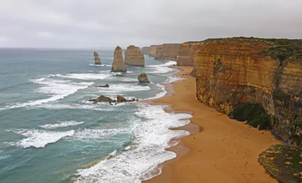 Waves crash along the sandy coastline of Victoria, Australia, showcasing the limestone stacks of The Twelve Apostles against a cloudy sky in this 4K Ultra HD wallpaper.