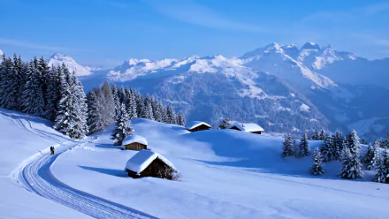 HD desktop wallpaper of a serene winter mountain landscape with snow-covered cottages and pine trees against a backdrop of majestic snowy peaks.