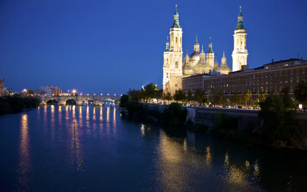 Night view of the Cathedral-Basilica of Our Lady of the Pillar along the Ebro River in Zaragoza, Spain, with an illuminated bridge and clear sky in the background.