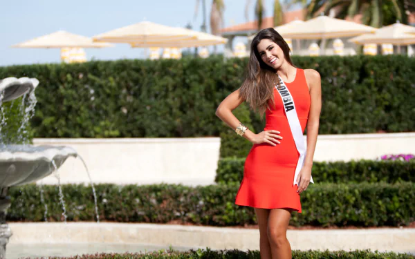 Miss Universe contestant Paulina Vega from Colombia poses elegantly in a vibrant red dress, showcasing a sparkling bracelet and a warm smile against a lush garden backdrop.