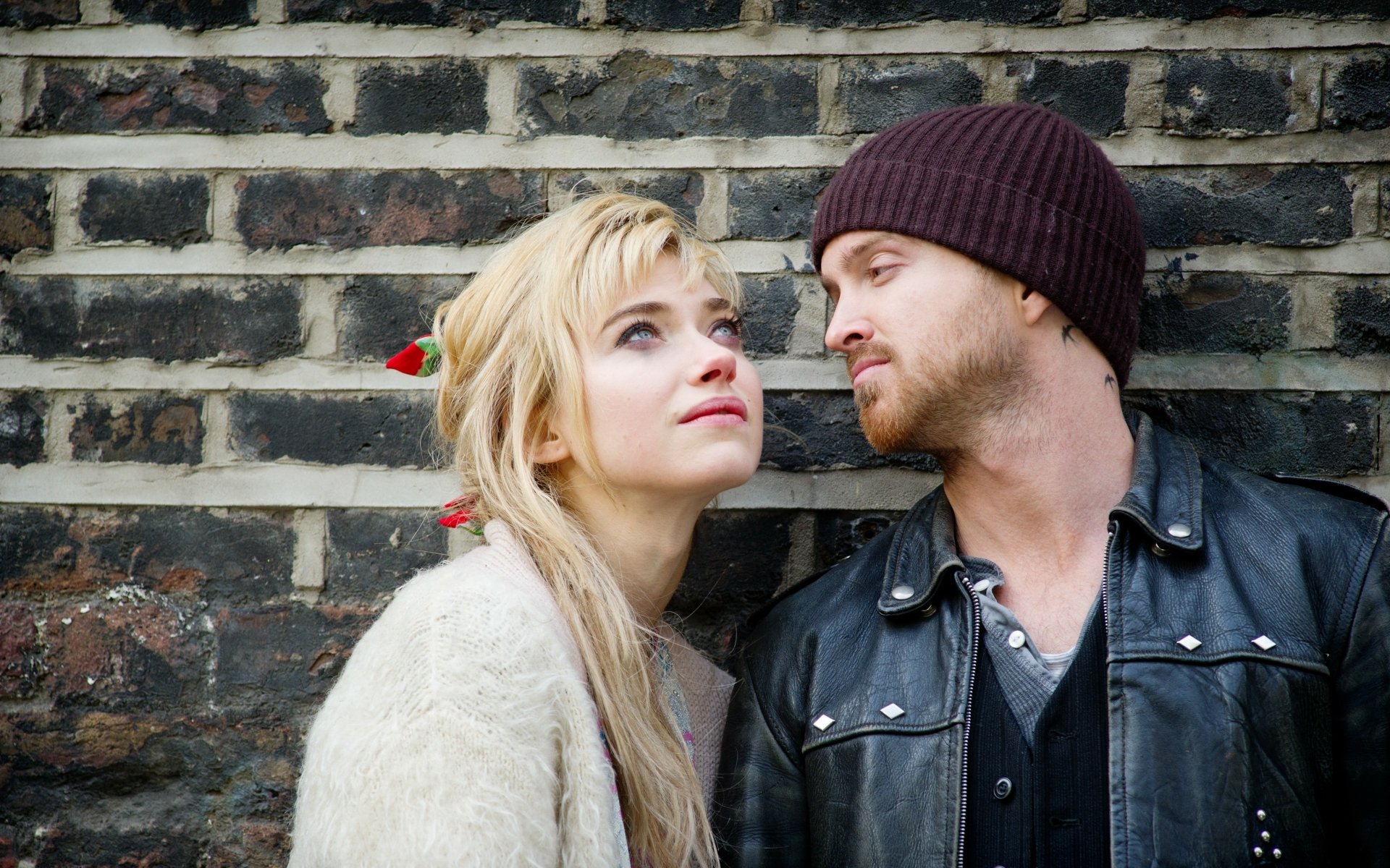 4K desktop wallpaper: blue-eyed blonde woman and bearded man in a knit cap lean against a brick wall, intimate movie still evoking A Long Way Down.
