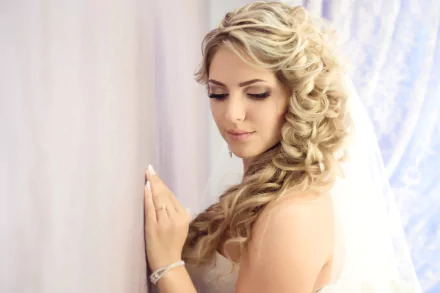 HD desktop wallpaper of a bride with curled hair wearing a wedding dress, softly posing by a light-colored wall.