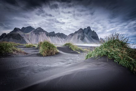 Vestrahorn Mountain rises dramatically behind black sand dunes with patches of green grass under a cloudy sky in this HD nature desktop wallpaper.