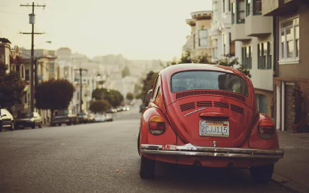HD wallpaper of a vintage red Volkswagen Beetle parked on a street lined with townhouses, evoking a nostalgic urban vibe.
