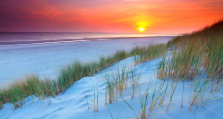 HD desktop wallpaper of a beach at sunset, featuring ocean waves, sandy dunes with grass, and vibrant natural colors.