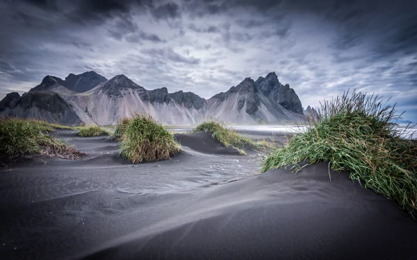 Vestrahorn Mountain rises dramatically behind black sand dunes with patches of green grass under a cloudy sky in this HD nature desktop wallpaper.