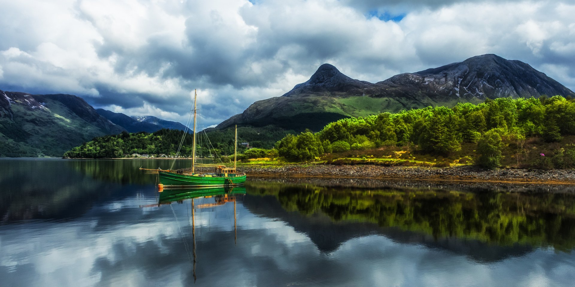 HD desktop wallpaper of a tranquil lake with boat reflections, surrounded by forested coast and towering mountains beneath a cloudy sky.