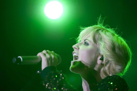 Blonde female singer mid-performance, gripping a microphone and singing under a bright green stage light — energetic live-music portrait as a 4K Ultra HD PC desktop wallpaper background
