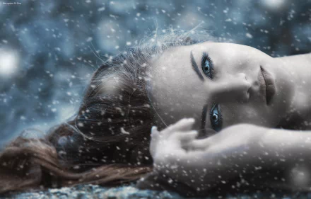 A high-definition desktop wallpaper featuring a woman with blue eyes, lying amidst falling snow, capturing an artistic and serene winter scene.